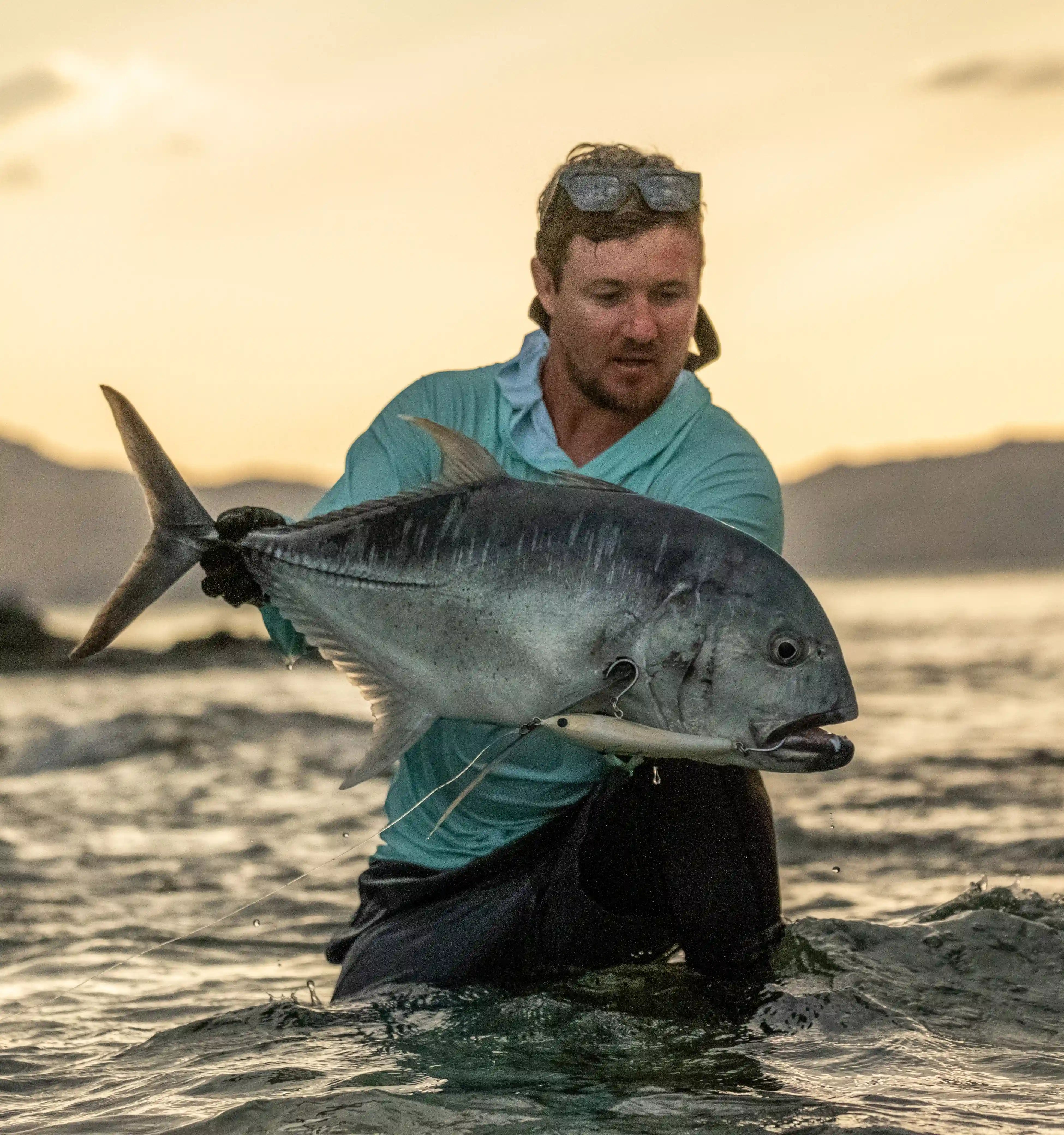 Adam Cavanagh holding giant trevally on the shore at sunset – Cavy Fishing mission