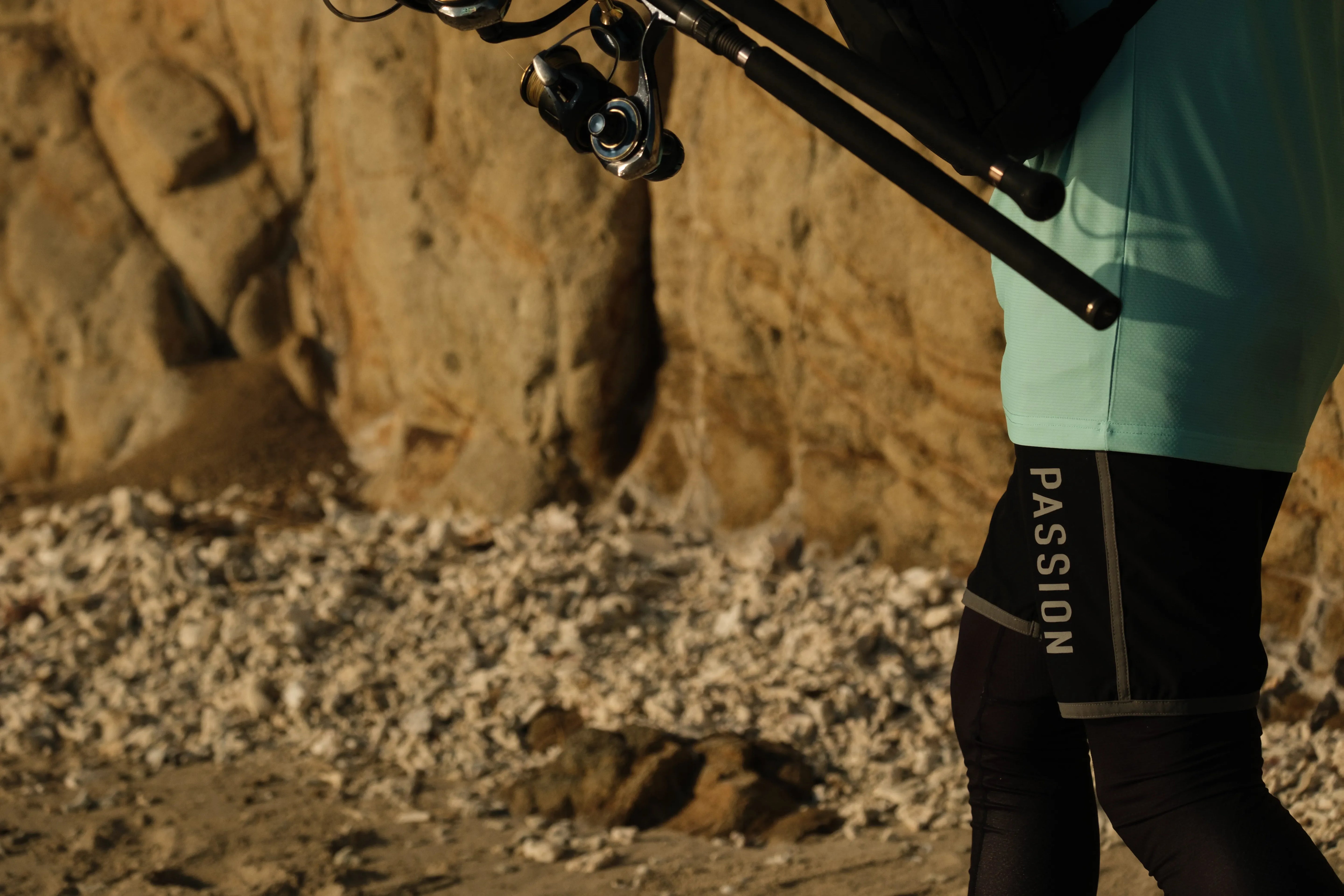 Angler wearing Cavy Fishing shorts holding fishing rods near rocky coastline, showing quick-dry saltwater fishing apparel tested in tropical conditions.
